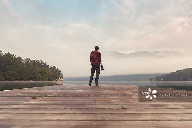 男人站在湖上码头，以天空为背景，手持相机的背影图片素材