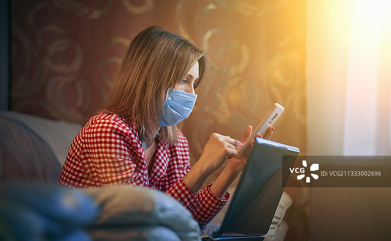 戴外科口罩的女人在家工作时使用笔记本电脑和智能手机图片素材