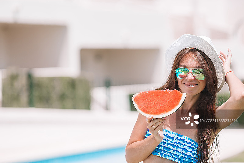 微笑的年轻女子拿着西瓜站在游泳池旁的肖像图片素材