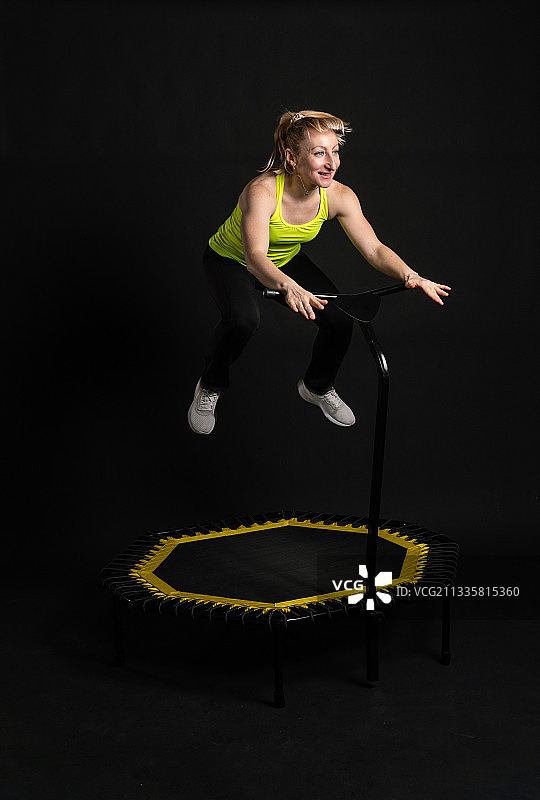 中年妇女穿着黄色T恤在黑色背景上使用健身蹦床图片素材