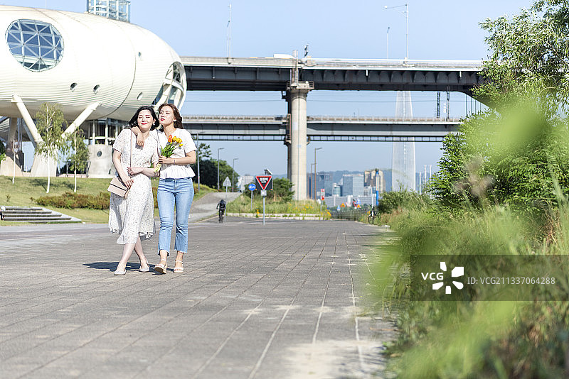 两名女子在公园沿河散步，手捧鲜花图片素材