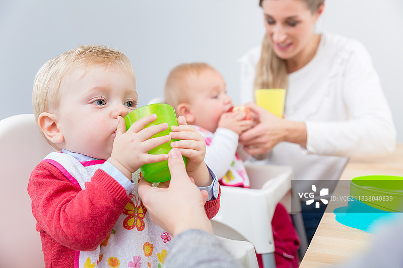 这是一个可爱的小男孩学习用塑料杯喝水的肖像，他坐在家里或现代婴儿日托中心的高椅子上，旁边是另一个婴儿图片素材