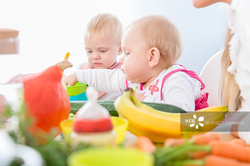 这是一张现代婴儿日托中心里一个可爱的蓝眼睛小女孩吃着健康的固体食物的照片，她和另一个婴儿一起坐在高椅子上图片素材