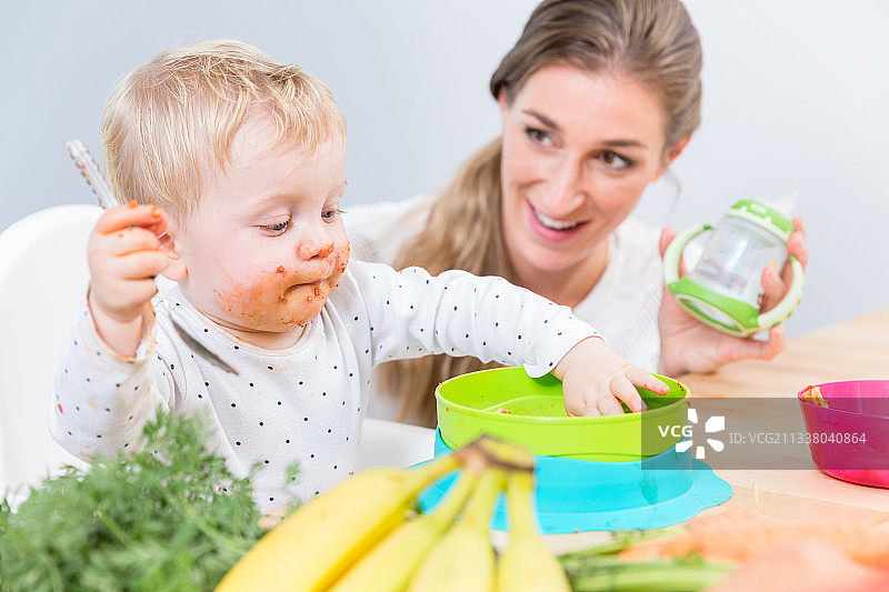 一个可爱和有趣的小女孩坐在高椅子上，用她的手从一个塑料碗吃固体食物旁边她奉献的母亲在家图片素材