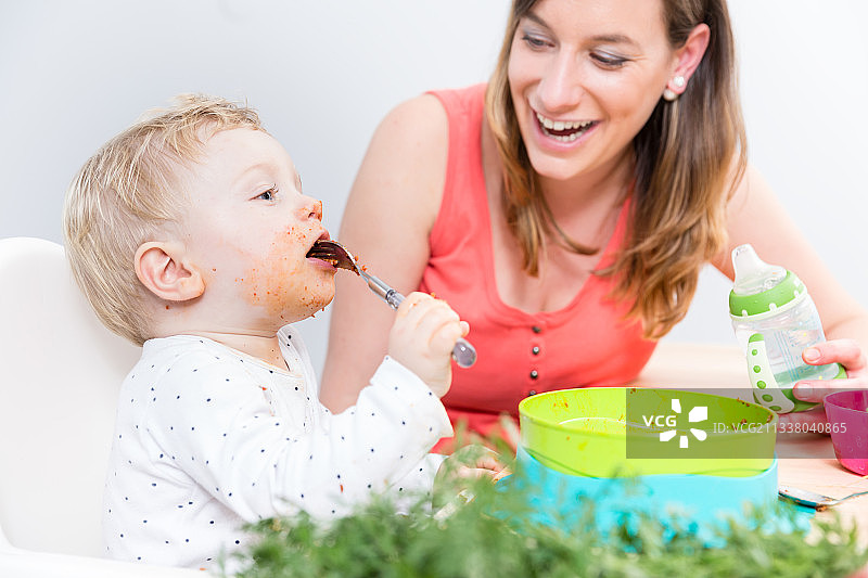 一个可爱和有趣的小女孩坐在高椅子上，用她的手从一个塑料碗吃固体食物旁边她奉献的母亲在家图片素材