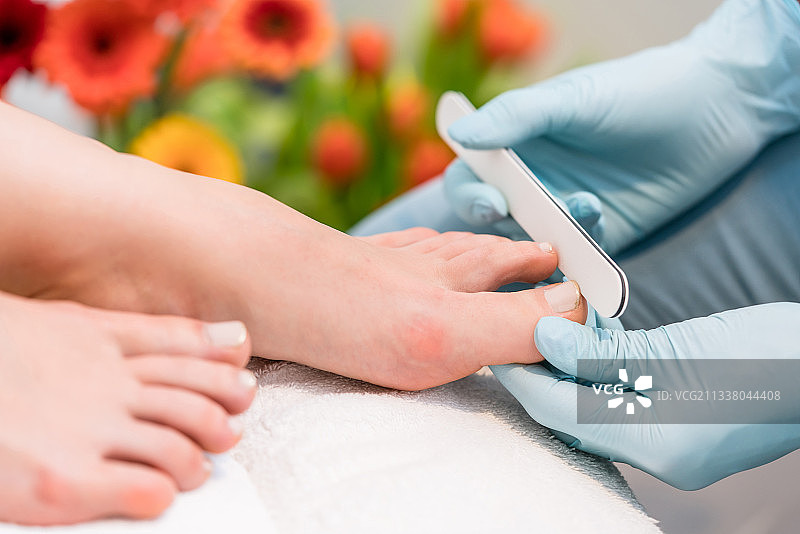 一个戴着外科手套的美甲师的手的特写，同时在一个现代美容沙龙的一个女性客户的脚趾甲边缘图片素材