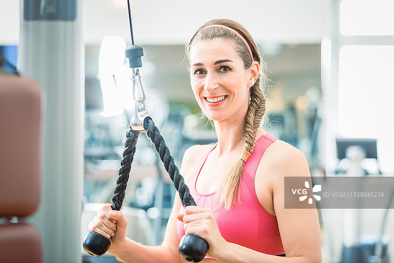 一个美丽的健康女人的肖像，微笑着看着镜头，而锻炼三头肌在绳子电缆机下推在健身房锻炼手臂图片素材