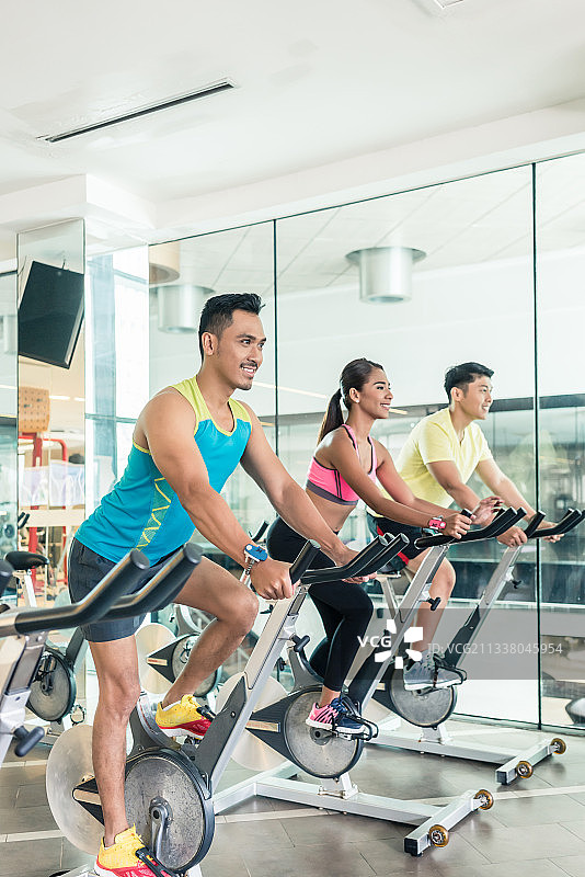 在一家现代健身俱乐部的HIIT室内自行车课上，帅气自信的年轻人微笑着骑自行车图片素材