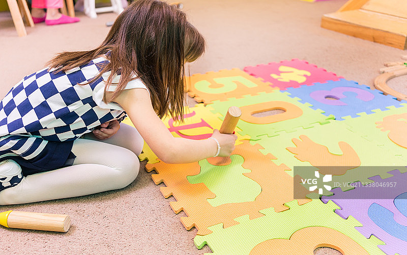 侧面的一个可爱的学前女孩拿着木制玩具积木，而玩一个彩色的拼图游戏垫与数字和符号在一个现代幼儿园的教室图片素材