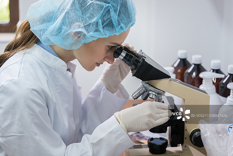 在化妆品工厂的化验室中，一名女化验师戴着无菌安全设备在显微镜下分析样本的侧视图图片素材
