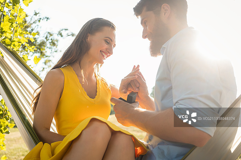 低角度的一个快乐的年轻人把订婚戒指在他的女朋友的手指上，接受他的建议，而坐在一起在一个阳光明媚的夏天的吊床上图片素材