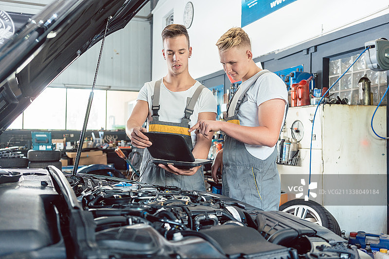 在一家现代汽车修理厂，有经验的汽车修理工在一个学徒旁边用笔记本电脑扫描和解释引擎错误代码图片素材