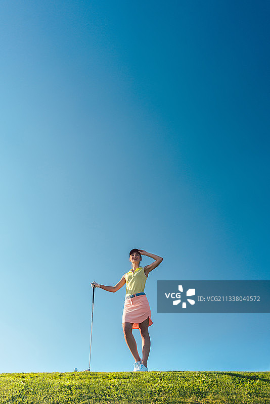 低角度的观点，一个健康和快乐的女人穿着高尔夫时髦的服装，而看着在一个专业高尔夫球场的绿色草地上练习图片素材