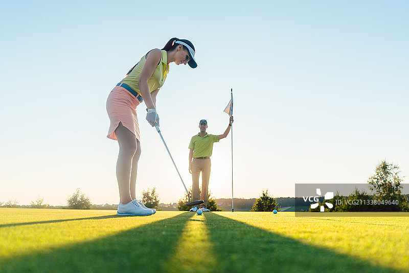 低角度侧面的一个有吸引力的健康的女人练习击球技术在高尔夫课上与一个经验丰富的职业球员在夏天图片素材