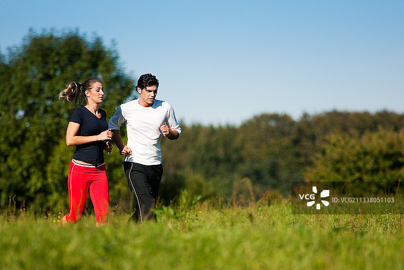 一对年轻的健身夫妇在户外做运动，在晴朗的蓝天下的绿色草地上慢跑图片素材