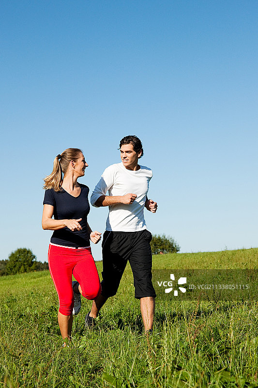 一对年轻的健身夫妇在户外做运动，在晴朗的蓝天下的绿色草地上慢跑图片素材