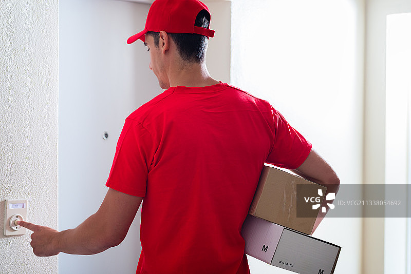 后视图的信使在红色制服携带纸箱按门铃图片素材