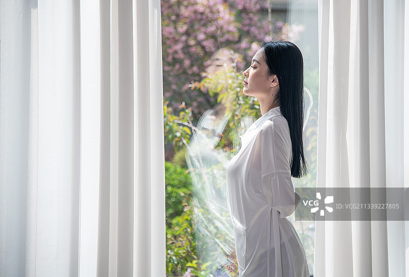 清晨，站在窗前刚起床的年轻女子，美丽优雅的青年女性。图片素材