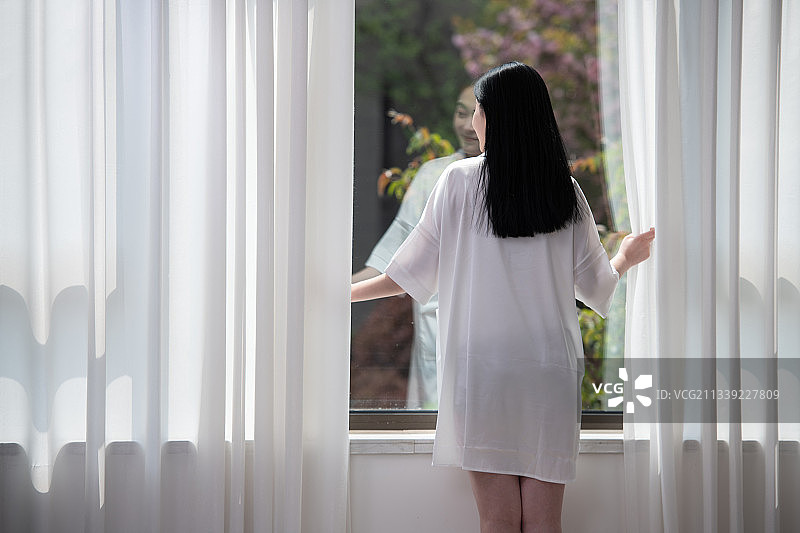 清晨，站在窗前刚起床的年轻女子，透过窗户往外看，美丽优雅的青年女性。图片素材