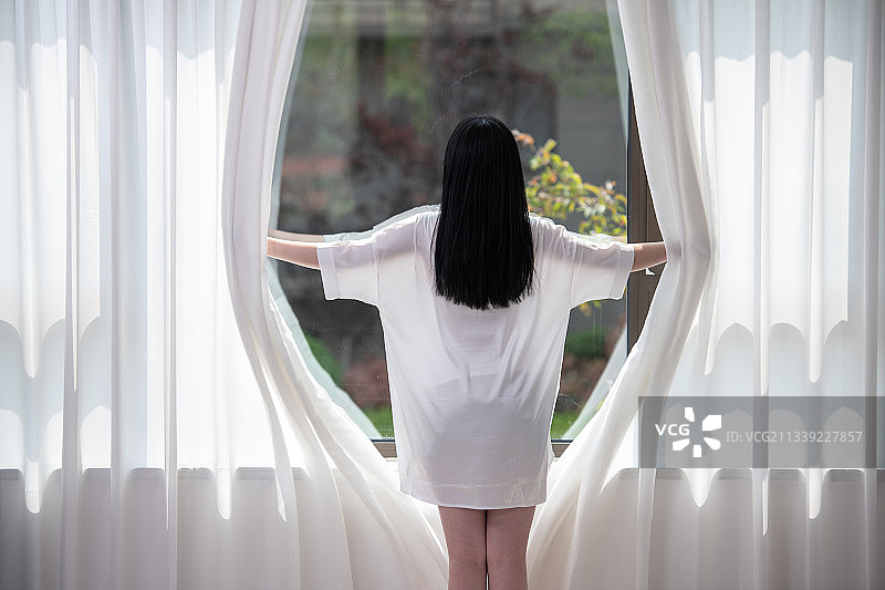 清晨，前刚起床的年轻女子打开窗帘，透过窗户往外看，美丽优雅的青年女性。图片素材
