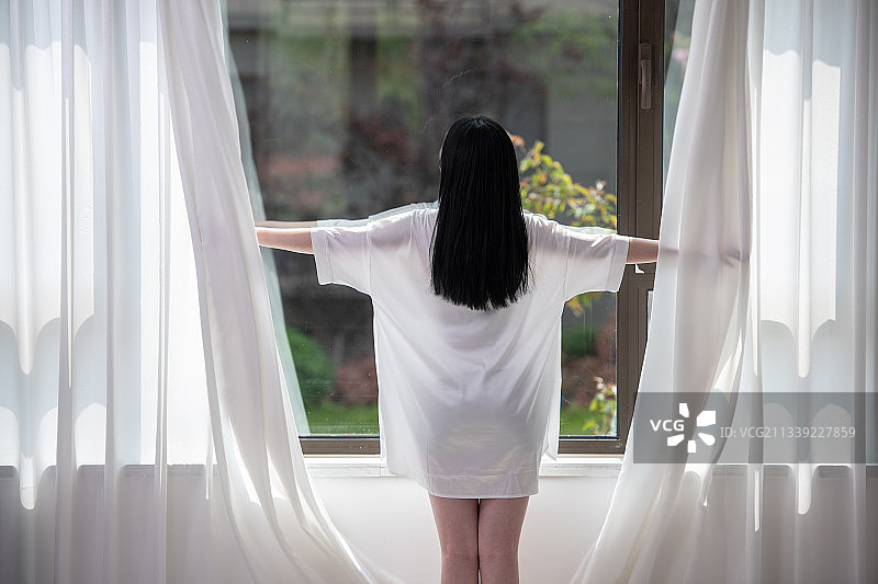 清晨，前刚起床的年轻女子打开窗帘，透过窗户往外看，美丽优雅的青年女性。图片素材