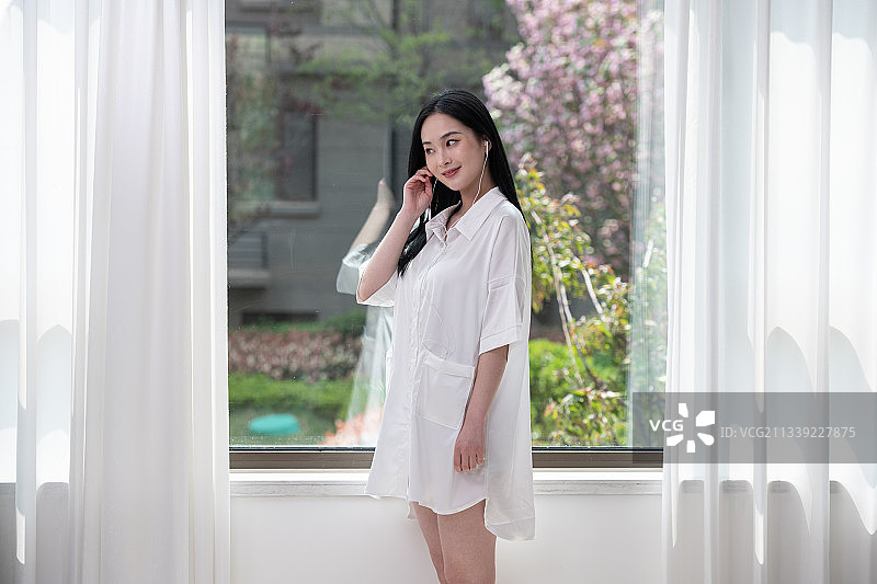 清晨，刚起床的年轻女子，站在窗前带上耳机听音乐，美丽优雅的青年女性。图片素材