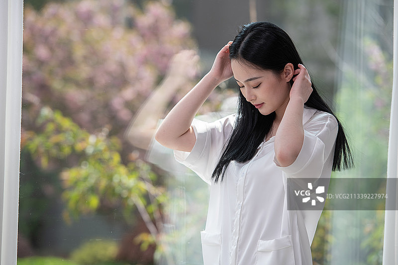 清晨，站在窗前刚起床的年轻女子，梳理黑色披肩长发，美丽优雅的青年女性。图片素材