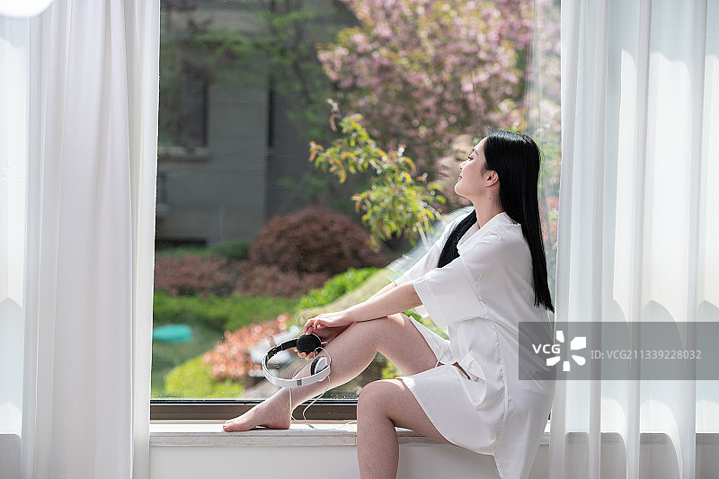 光着脚坐在窗前，拿着耳机，美丽优雅的青年女性。图片素材