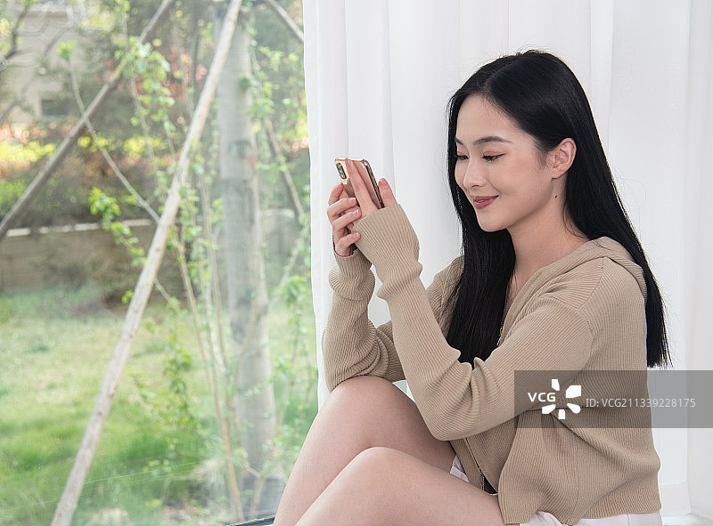 阳光明媚，坐在窗前使用移动电话的年轻女子，美丽优雅的青年女性。图片素材
