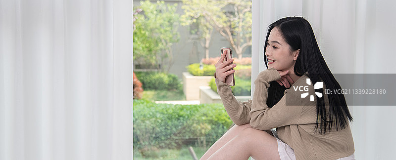 阳光明媚，坐在窗前使用移动电话的年轻女子，美丽优雅的青年女性。图片素材