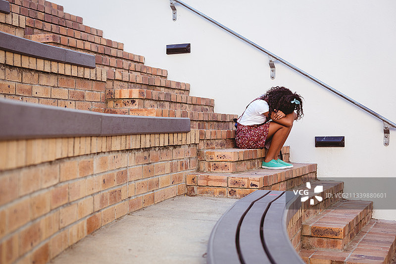 不快乐的非裔美国女学生坐在学校操场的台阶上，带着书包图片素材