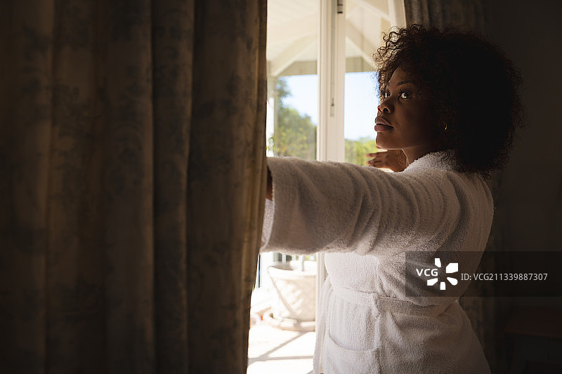 在阳光明媚的窗边拉开窗帘、向远处眺望的非裔美国妇女图片素材