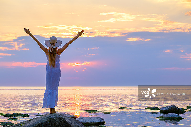 日落时分，女子在海滩岩石上张开双臂，仰望天空图片素材