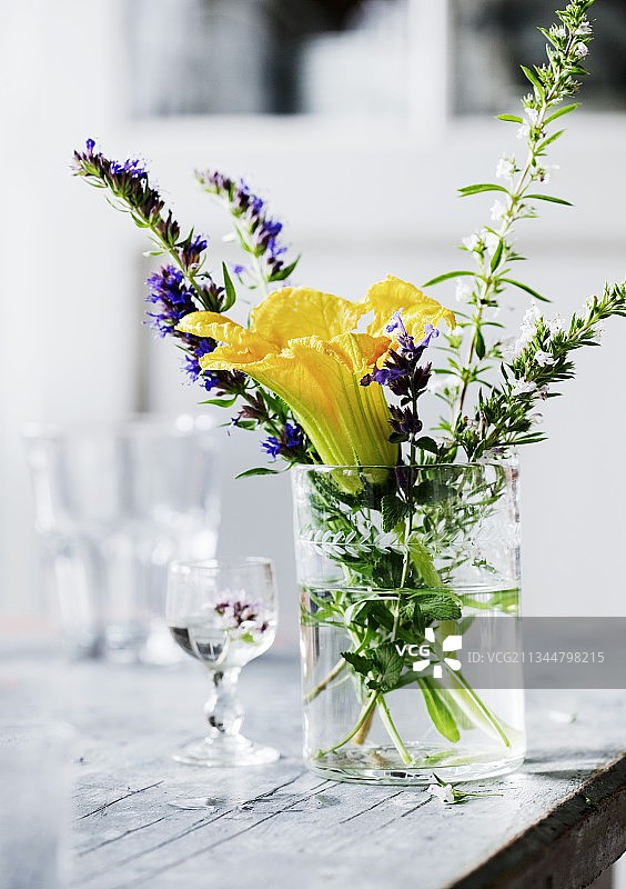 旧桌子上装在水杯里的黄色小胡瓜花和开花草药图片素材