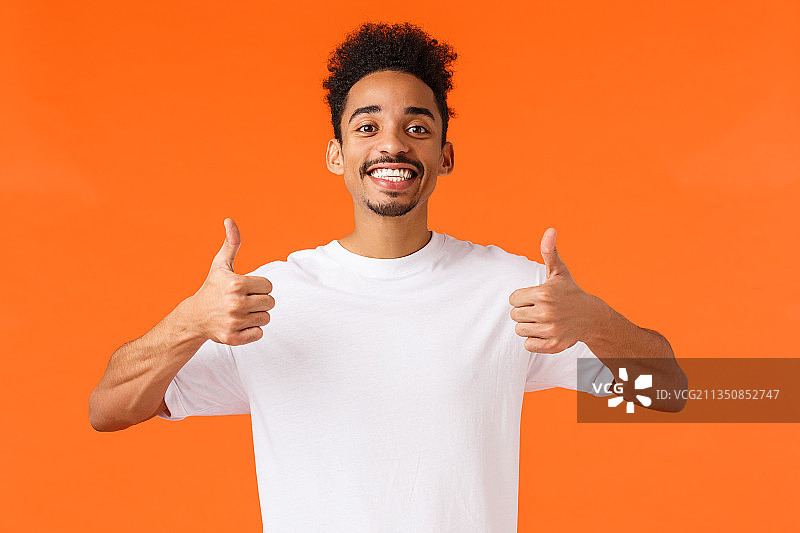 在橙色背景前竖起大拇指的微笑青年人像图片素材