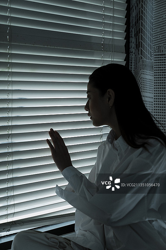 长镜头的长发女人在房间里透过紧闭的窗户向外看，摸了摸窗户，她想出去。图片素材