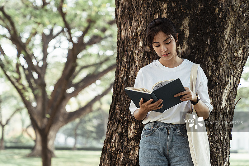 穿白t恤的可爱女孩在院子里的大树旁看书。图片素材