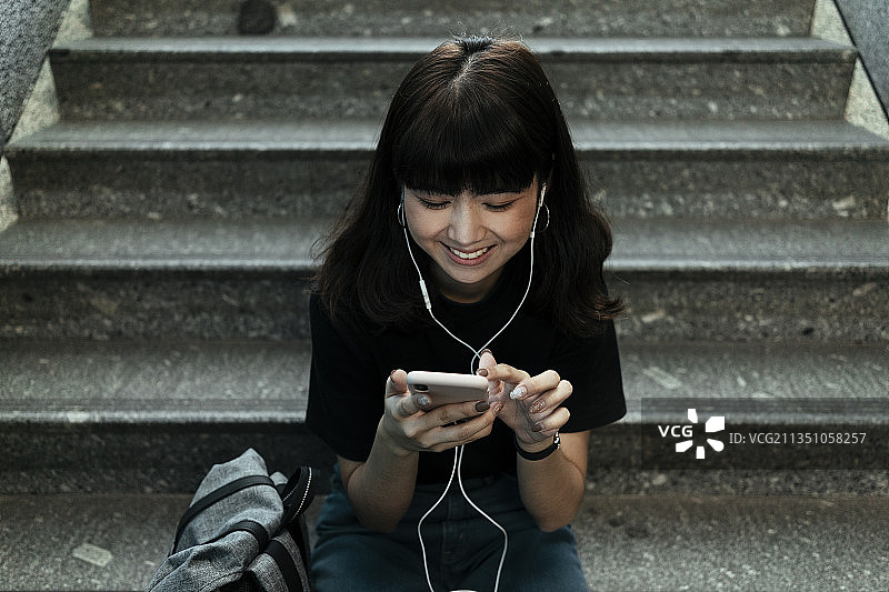 亚洲泰国背包客女人喜欢听音乐从智能手机耳机，坐在楼梯上。图片素材