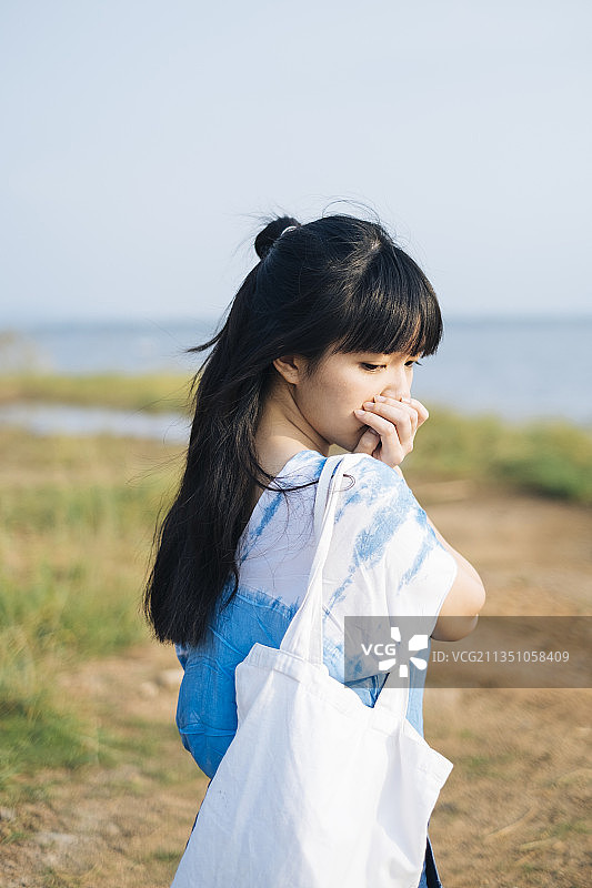 中景，黑色半扎头发的女孩穿着白色和蓝色的扎染，站在海边的草原上，拿着帆布包，闭着嘴。图片素材