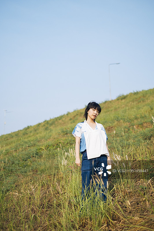 黑色半扎头发的女孩穿着白色和蓝色的扎染领带站在草原上，脖子上挂着帆布包。图片素材