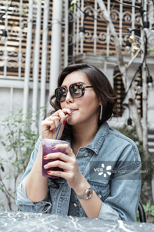 亚洲泰国女人穿着牛仔夹克，戴着太阳镜，在咖啡馆用吸管喝塑料杯里的葡萄汁。图片素材