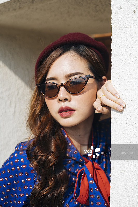 黑发卷发的女人穿着蓝色和红色的复古连衣裙站在大楼的白色框架前把头固定在框架上，靠近点。图片素材