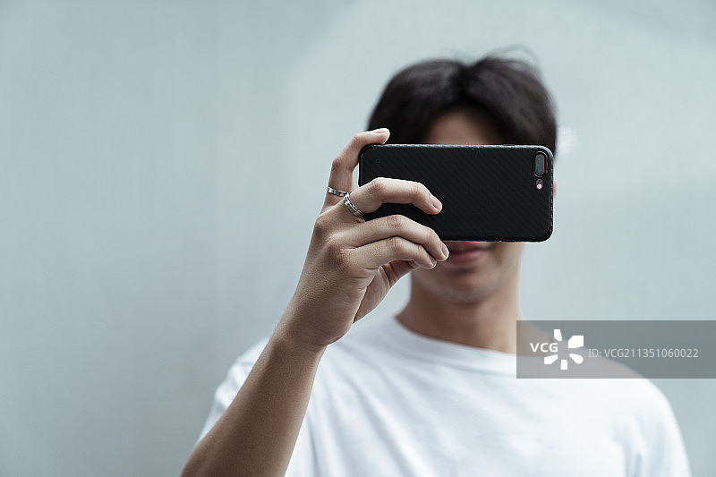 黑头发的家伙穿着白色t恤站在墙前，举起手机遮住了脸。图片素材