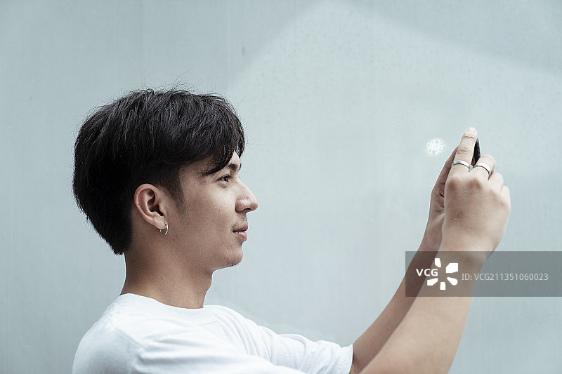 黑头发穿着白t恤的家伙站在墙前，举起智能手机拍照。图片素材