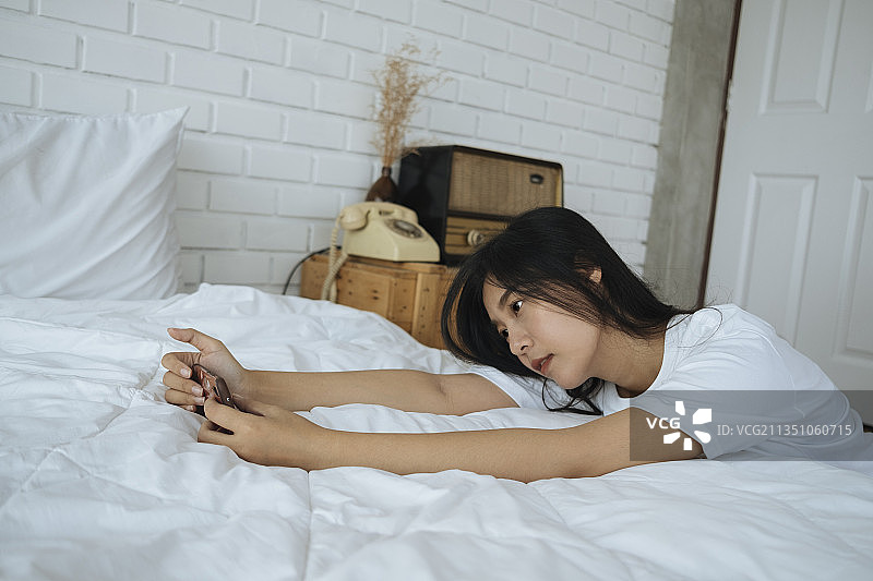 可爱的亚洲女人玩手机，和朋友聊天，在facebook上工作。白色的卧室。图片素材