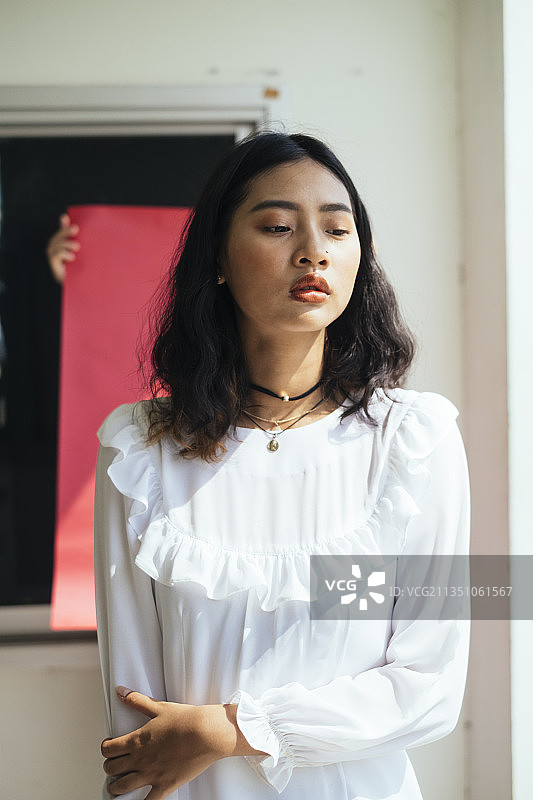 黑色卷发的长发女孩身穿白色连衣裙站在红纸前的灯光下。图片素材