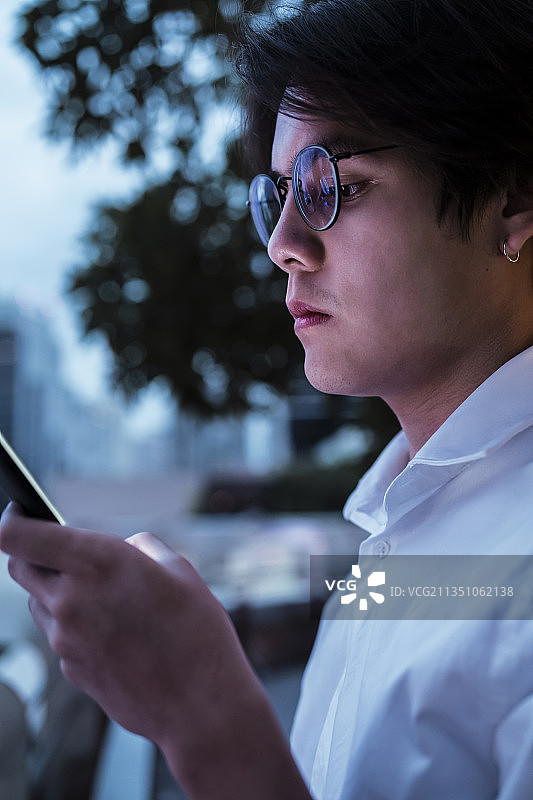身穿白衬衫、戴着眼镜的黑发男子站在屋顶上，看着他的智能手机读着朋友发来的信息，背景是城市的夜景。图片素材