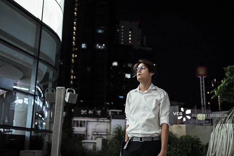 黑头发的家伙，穿着白衬衫，戴着眼镜，在夜里走过首都中心的LED广告牌购物中心。图片素材
