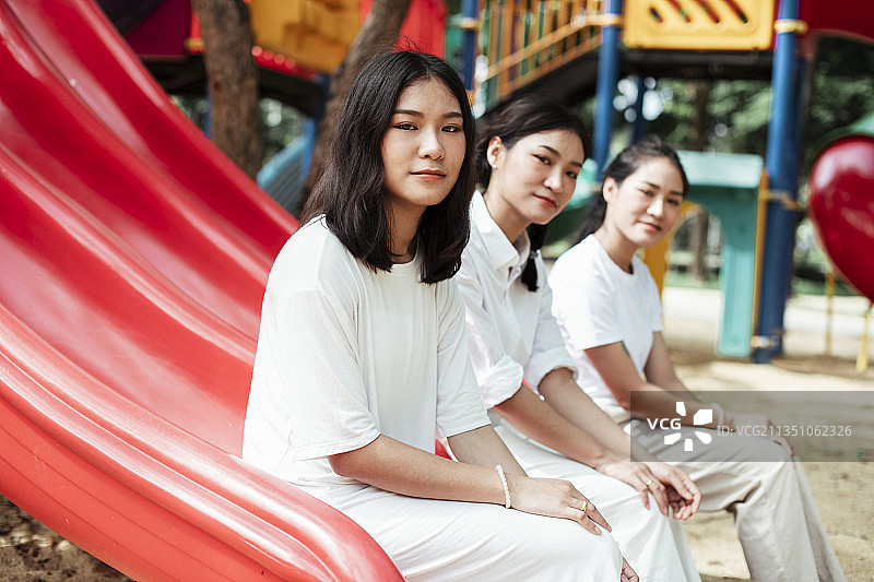 三个双胞胎姐妹坐在操场的红色滑块的末端，看镜头。图片素材