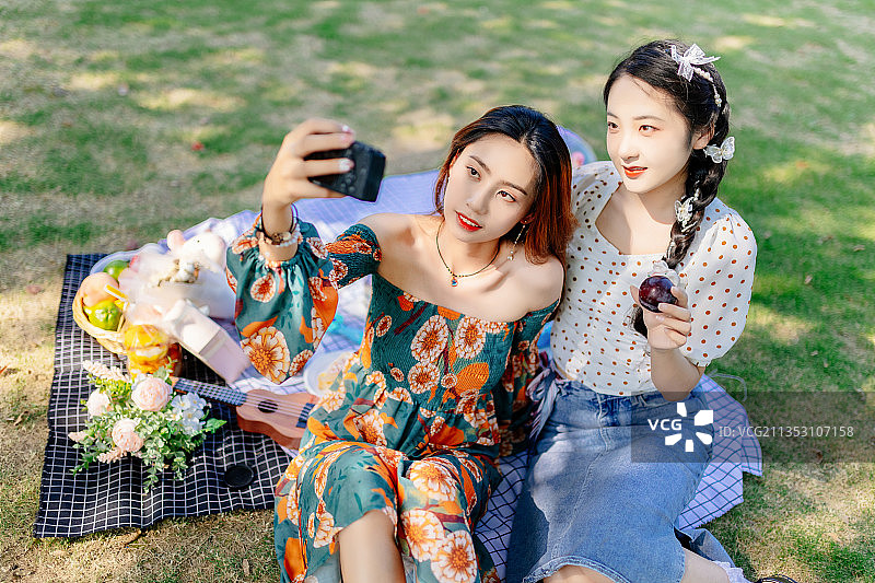 两个亚洲青年美女闺蜜在户外野餐垫上野餐用数码相机给对方拍照图片素材